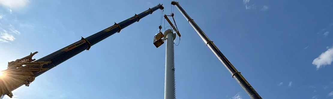 blauer Himmel, sonne scheint am linken Bildrand rein, zwei Kranspitzen sind neben einem Stahlkamin links und rechts zu sehen, Montagearbeiten
